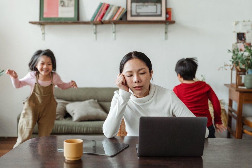 Mother sitting at table staring at laptop as son and daughter rambunctiously play in living room